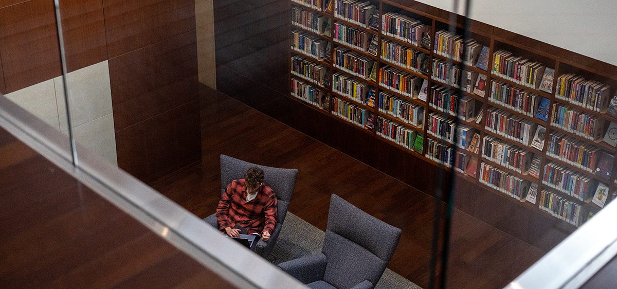 A student sitting in a chair in Hannon Library, with a large bookshelf full of books behind them, with the camera on a balcony above aiming down at the student.